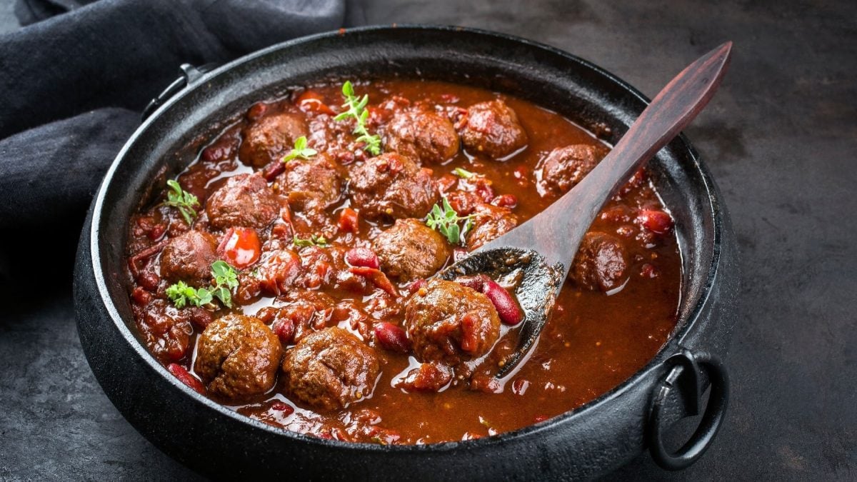 Ein schwarzer Topf mit den Hackbällchen in einer Tomatensauce mit Bohnen und PAprika auf schwarzem Untergrund.