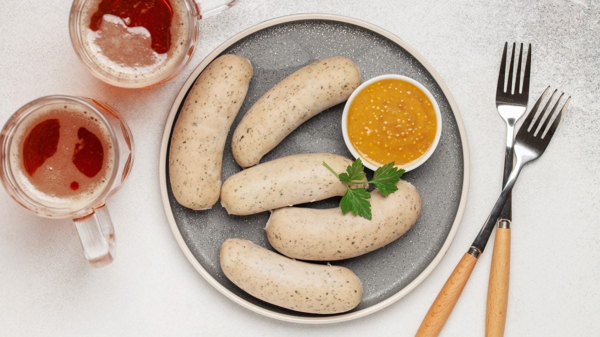 Fünf vegane Weißwürste mit einer Schale süßem Senf auf einem Teller, daneben zwei Gabeln und zwei Biergläser. Draufsicht.