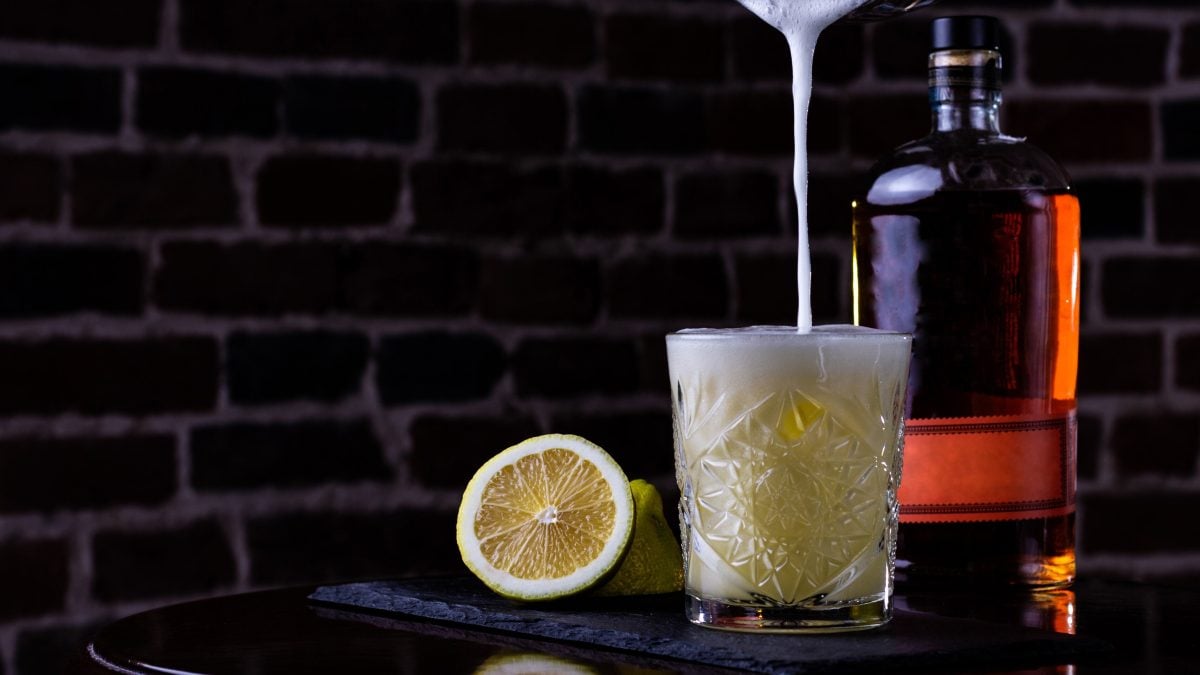 Whiskey Sour oder Boston Sour beim Eingießen in Old Fashioned Glas vor auf Holztisch Backsteinwand. Daneben Zitronen und Whiskeyflasche. Forntalansicht.