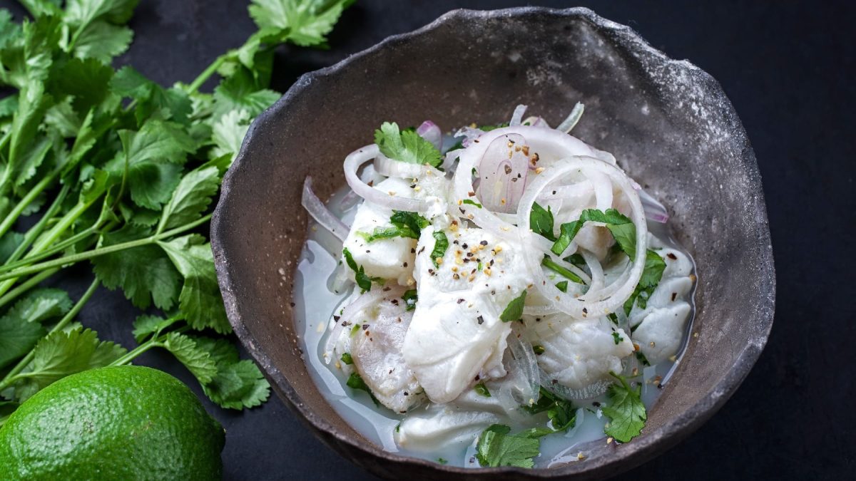 Wolfsbarsch-Ceviche auf einem grauen Teller und dunklem Untergrund. Neben dem teller ein Bund Koriander und eine Limette.
