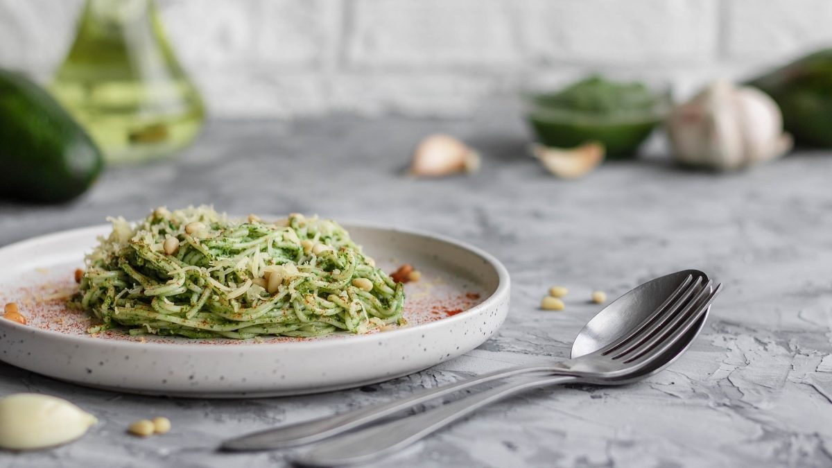 Ein Teller Zucchini-Avocado-Pasta mit Pinienkernen, daneben liegt Besteck und Knoblauch.