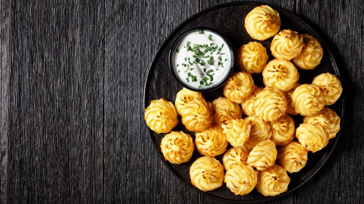 Ein schwarzer Teller mit Herzoginkartoffeln neben einem Dip auf dunklem Hintergrund.