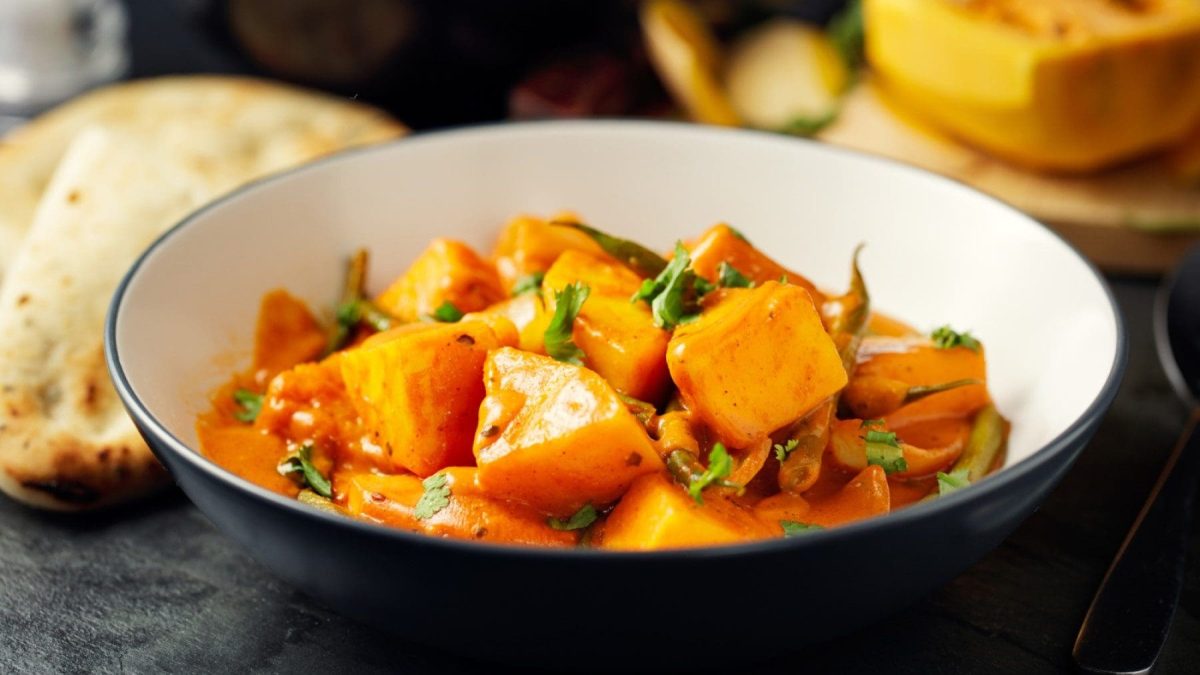 Tiefer Teller mit Kartoffel-Curry mit Kürbis auf schwarzem Untergrund, im Hintergrund ist Brot Kürbis zu sehen.