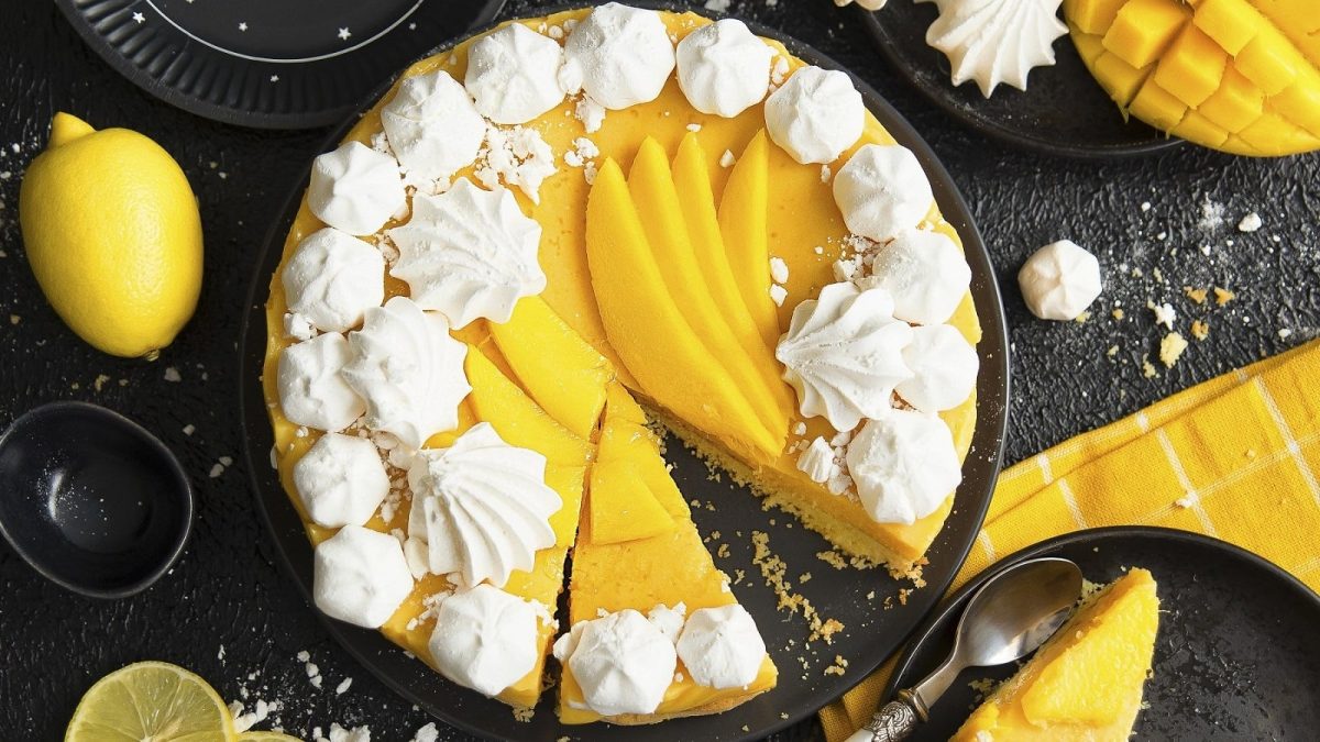 Auf einem Kuchenteller steht ein Mango-Käsekuchen mit Baisertropfer. Er ist mit frischer Mango belegt.