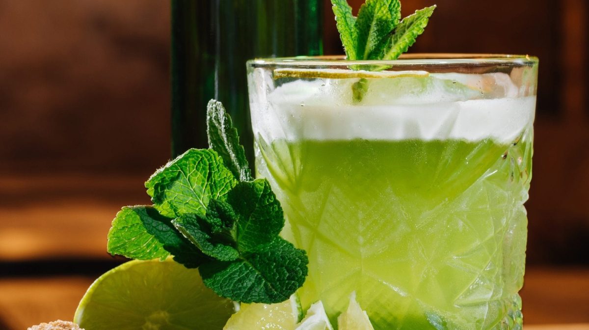 Absinth Sour auf Holztheke. Danaben Minze und Limetten. Im Hintergrund eine Flasche Absinth. Fronatlansicht.