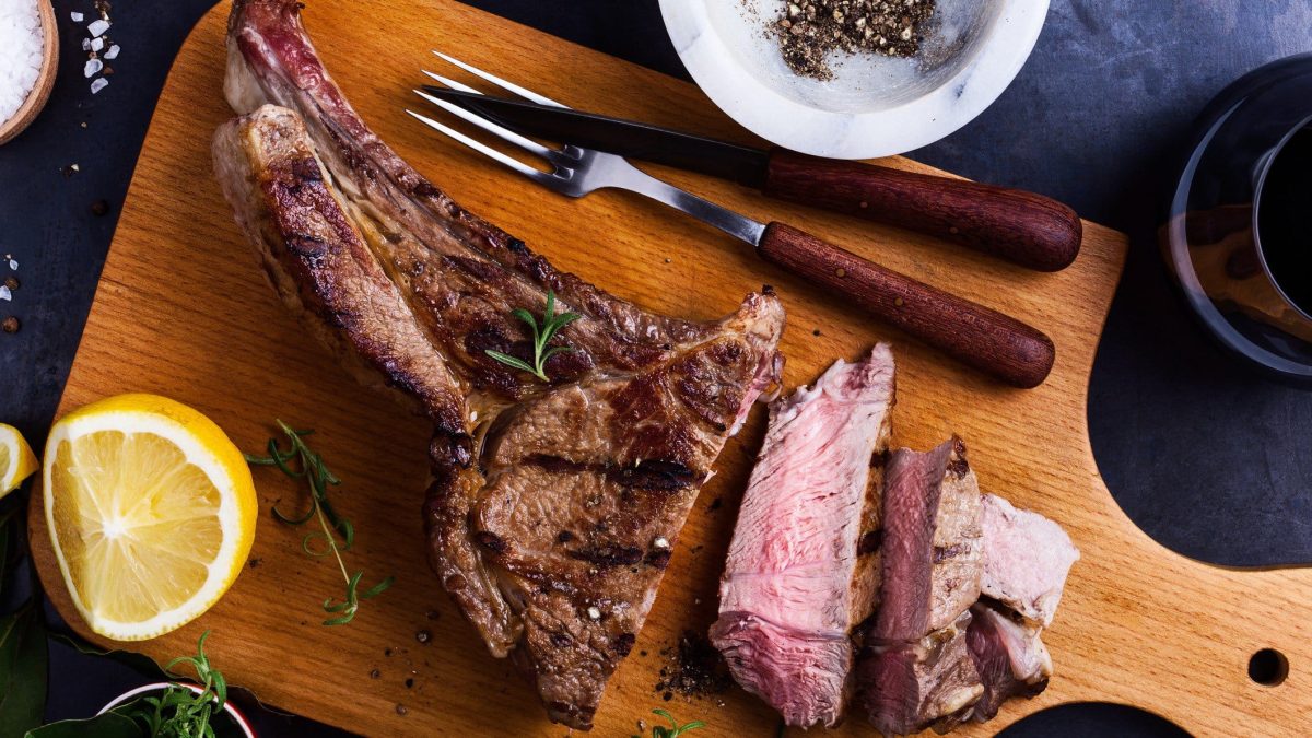 Anleitung zum Rückwährtsgaren für Entrecote Double am Knochen. Aufgeschnittenes Steak auf Holzbrett mit Besteck. Daneben eine Zitrone, Kräuter und je eine Schale mit schwarzem Pfeffer und grobem Salz. Draufsicht.