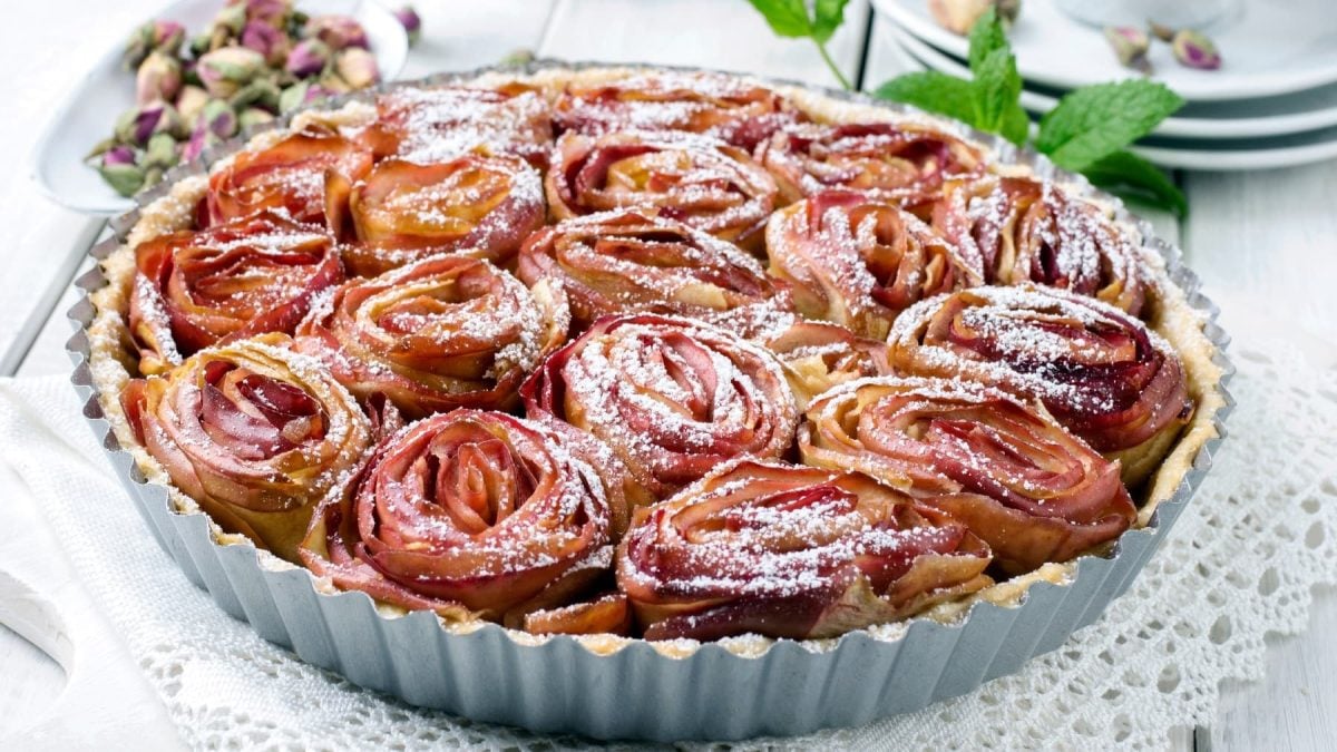 Eine silberne Springform mit der Apfelrosen Tarte auf hellem Untergrund. Daneben ein Zweig Minze und eine Schale mit Blüten.