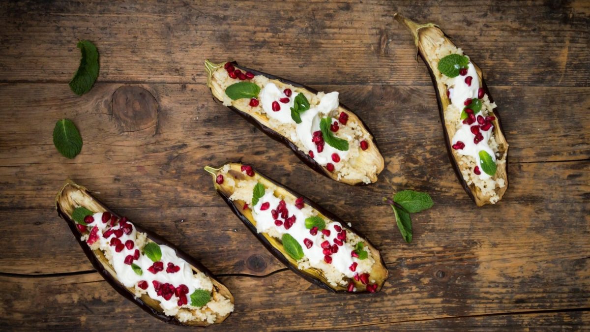 Gefüllte Aubergine mit Couscous, Granatapfel und Minzjoghurt auf Holz. Minzblätter als Deko im Bild verteilt. Draufsicht.