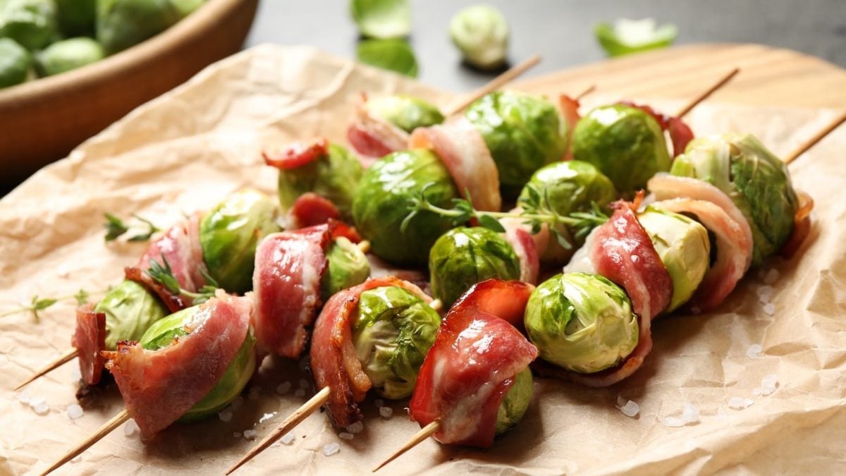 Frontal: vier Spieße, auf denen abwechselnd rosenkohl und bacon draufgewickelt sind, liegen auf einem holzbrett.