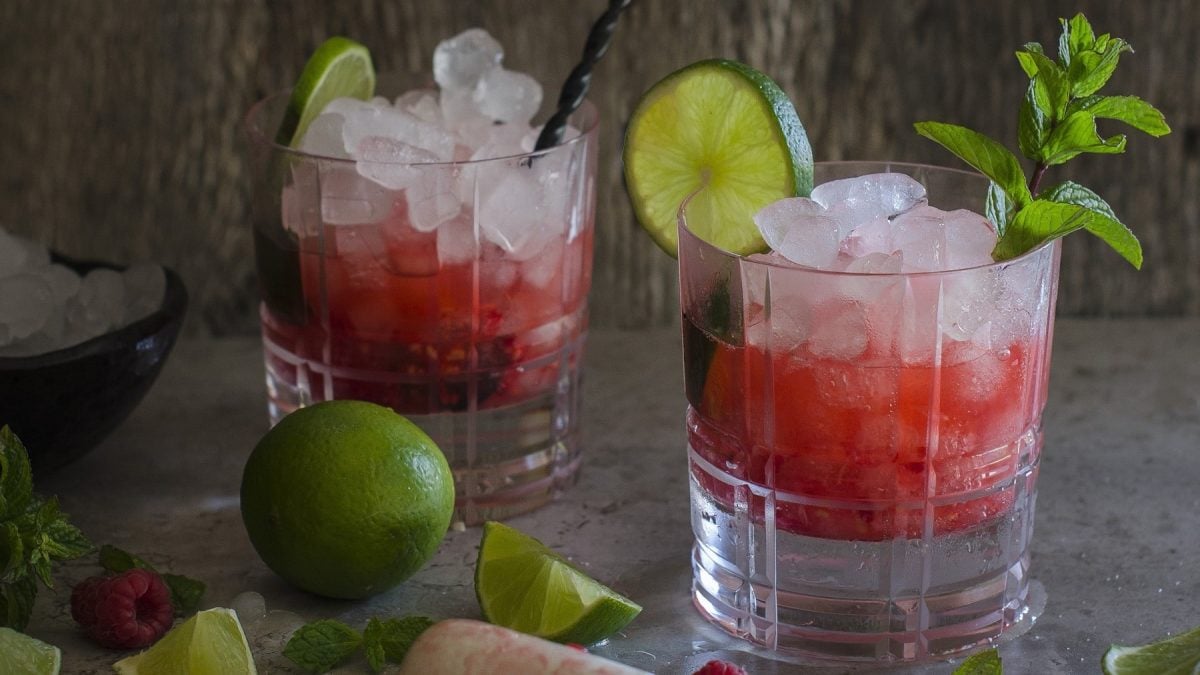 Zwei Gläser Caipiroska mit Himbeeren mit Minz garniert, neben Limetten vor grauem Hintergrund.
