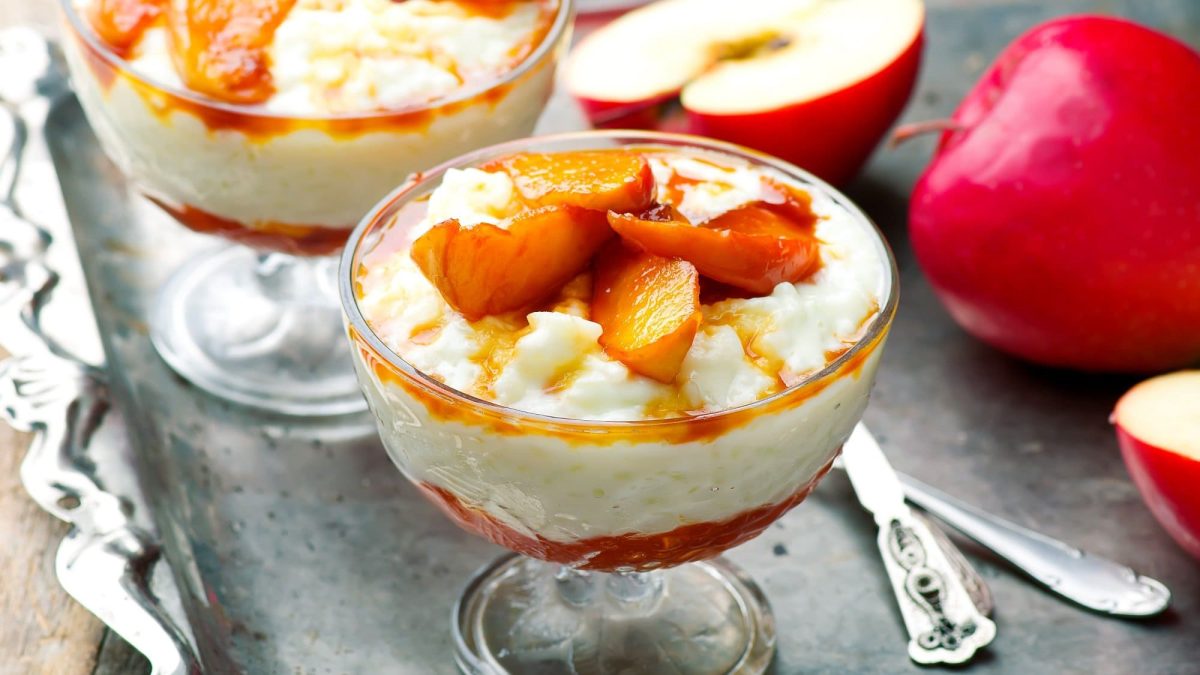 Zwei Dessertgläser Chai-Milchreis mit Zimtäpfeln, daneben liegt ein halbierter Apfel.