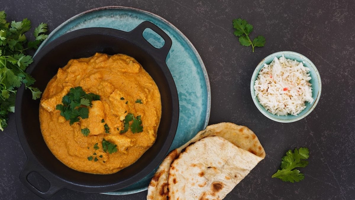 Eine schwarze Schüssel mit Chicken Korma auf einem hellblauen Teller neben Naan, Reis und frischem Koriander.