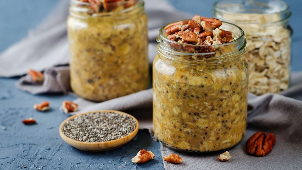 Zwei Gläser mit Kürbis Overnight Oats auf einem blauen Tisch neben Chiasamen und Nüssen.