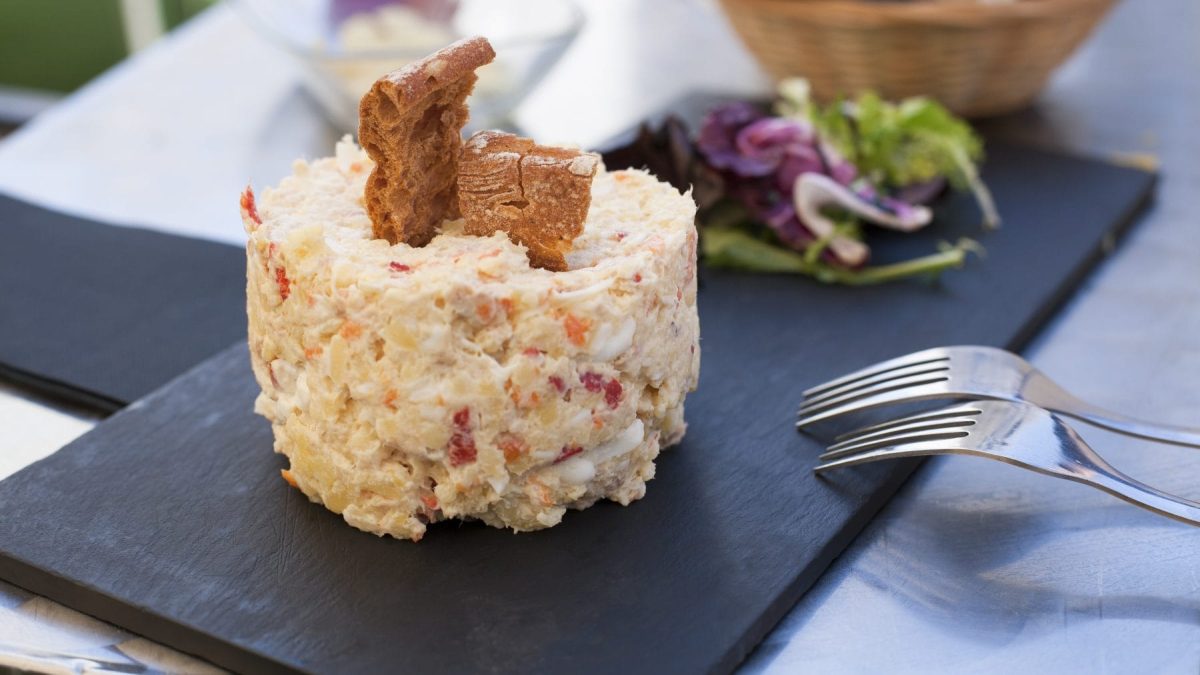 Ensalada Rusa angerichtet mit Salat und Brotchips auf Schieferplatte. Daneben zwei Gabeln. Frontalansicht.