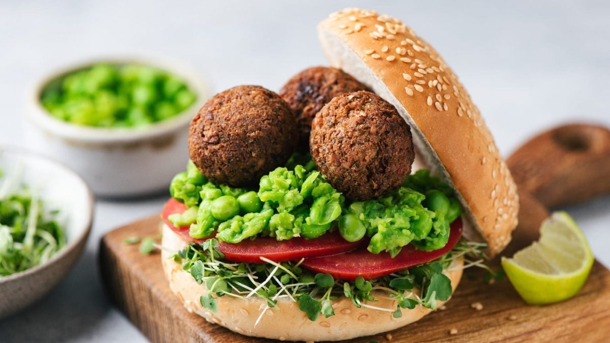 Ein aufgeklapptes Burgerbrötchen mit drei Falafelkugeln und etwas Erbsen-Guacamole auf einem kleinen Holzschneidebrett..