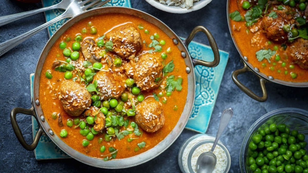 Fleischbällchen in Currysauce mit grünen Erbsen in einem Topf neben Zutaten vor blauem Hintergrund.