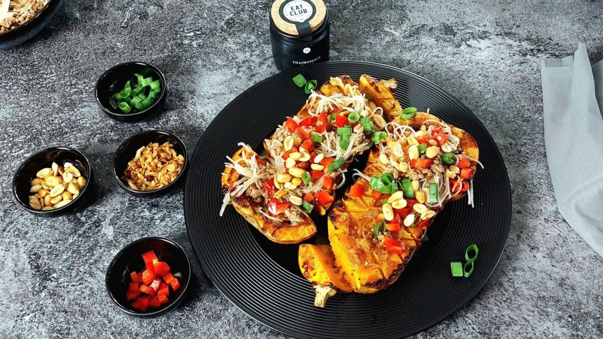 Gebackener Butternut-Kürbis Teriyaki mit Glasnudelsalat und Toppings auf schwarzem Teller und grauem Untergrund. Daneben Schalen mit Paprika, Erdnüssen, Röstzwiebelm und Lauch. Draufsicht.