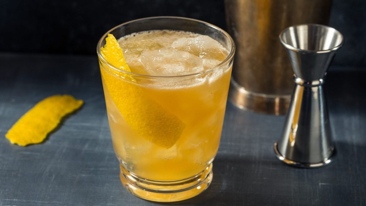 Gold Rush Bourbon auf grauem Untergrund. Im Hintergrund Barmaß, Shaker und Zitronenschale. Frontalansicht.