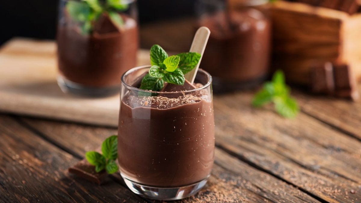 Frontalsicht: Zwei Gläser mit einem dunklen Pudding stehen auf einem Untergrund aus Holz. Auf dem Pudding ist ein Blatt Minze.