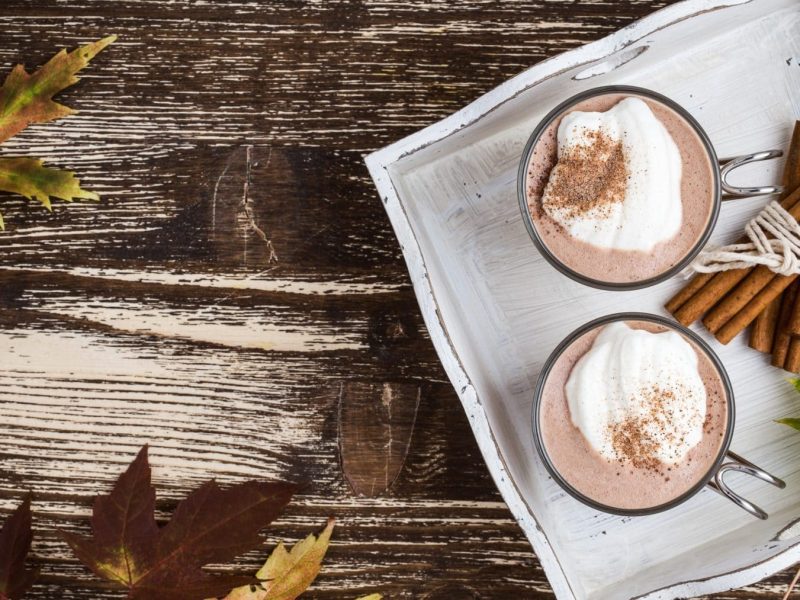 Heiße Schokolade mit Nougat und Sahne auf weißem Teller mit Zimtstangen und Blatt. Daneben herbstliche Blätter. Draufsicht.