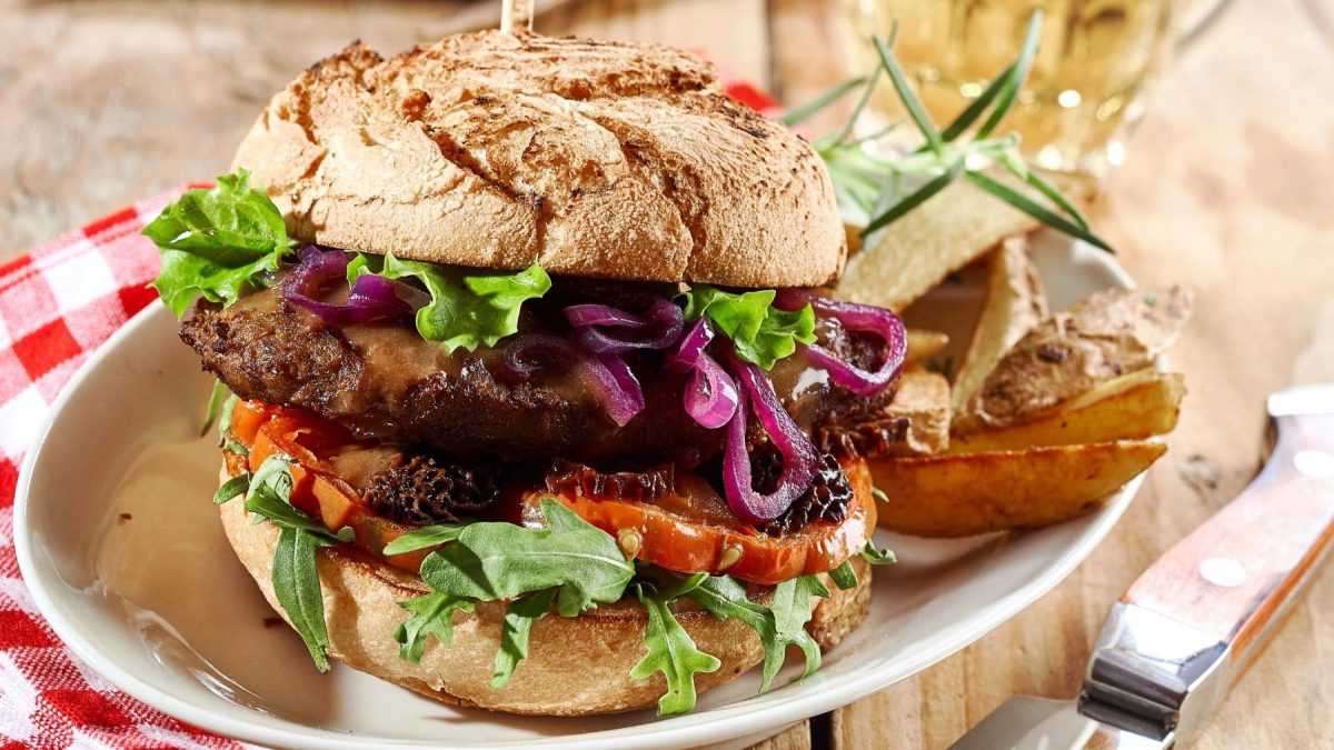 Wildburger vom Hirsch im Malzbrötchen auf Teller mit Kartoffelecken. Im Hintergrund ein Bierglas, rot-weiße Stoffserviette. Vorne Messer mit Holzgriff. Frontalansicht.