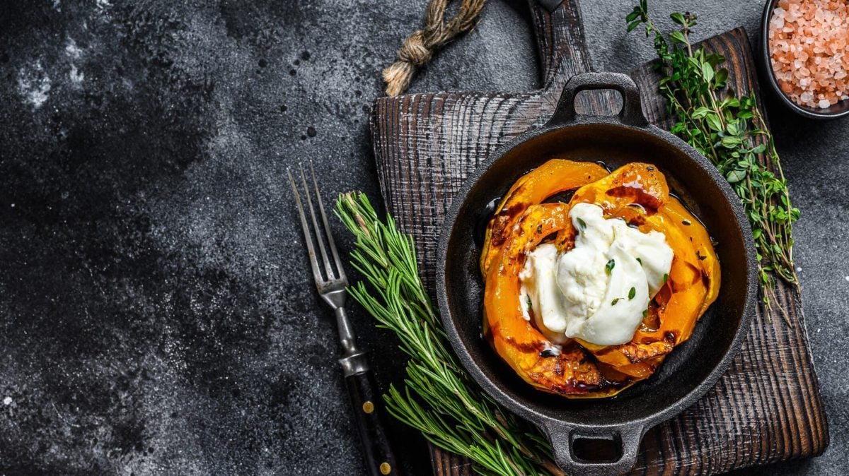 Gebackene Hokkaidospalten mit Burrata in schwarzer Auflaufform auf grauem Tuch. Daneben Kräuter und eine Schale mit pinkem Salz. Draufsicht.