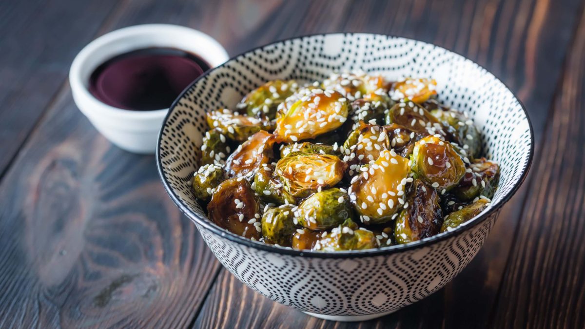 Japanischer Rosenkohl Gomasio Teriyaki in japanischer Schale auf Holz. Dahinter Schale mit Sojasauce. Draufsicht.