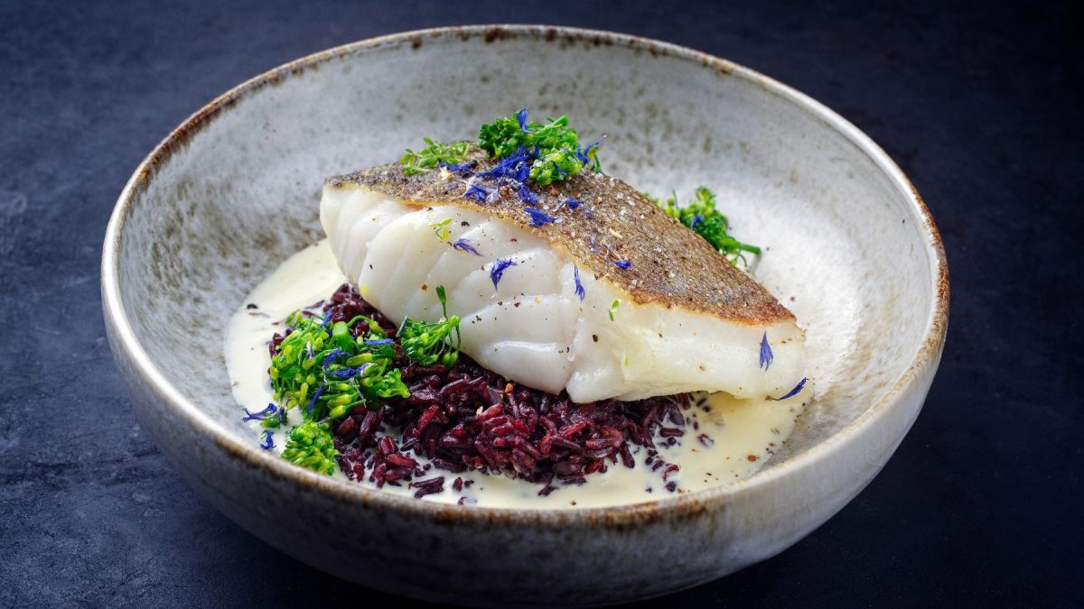 Ein grauer Teller mit schwarzem Reis und darauf ein Stück Kabeljau. Als Saucenspiegel eine helle Sauce udn alles dekoriert mit etwas Brokkoli und blauen Kornblumenblüten.
