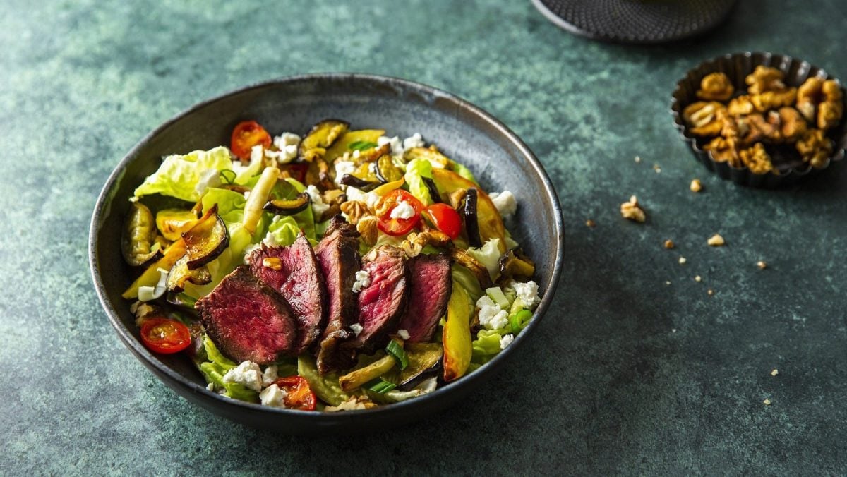 Kaffeemariniertes Steak auf Grillgemüse-Salat mit Nüssen und Feta in einem schwarzen Teller vor dunklem Hintergrund.