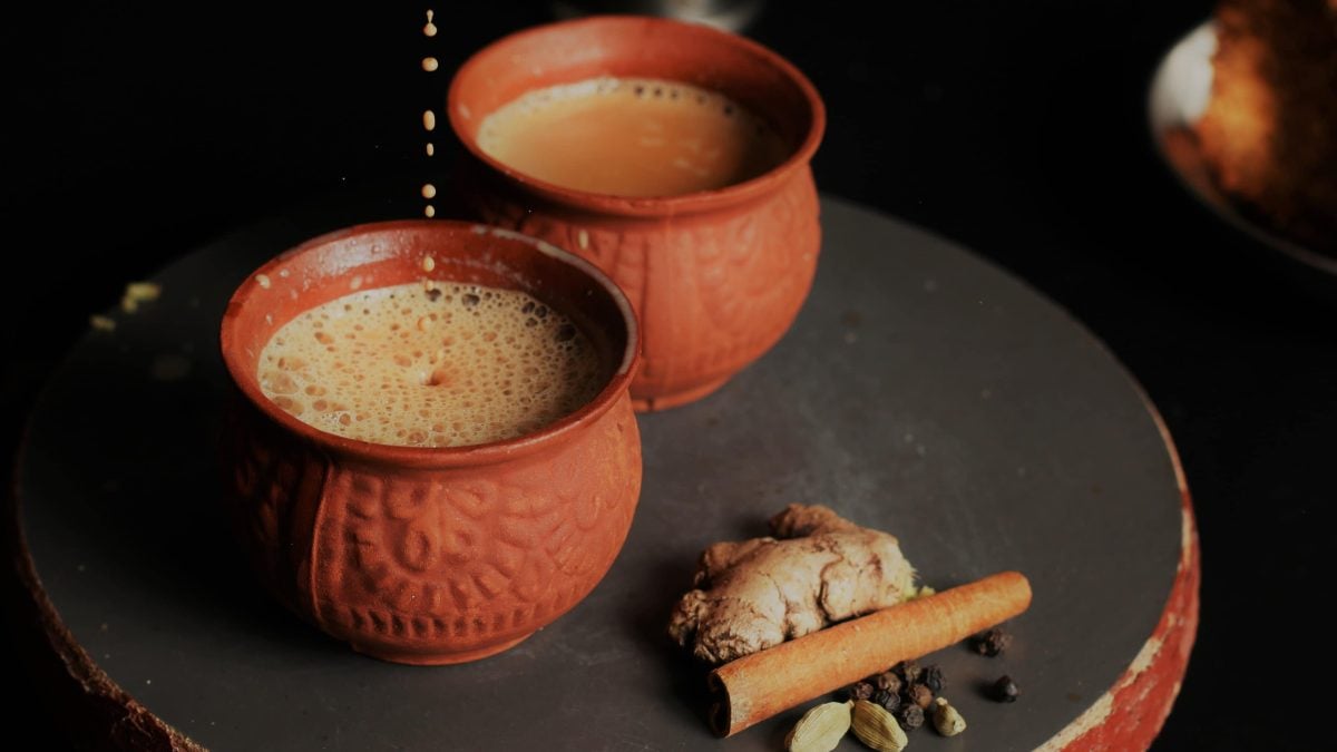 Masala Chai in zwei Tontassen auf dunkler Platte. Daneben Chai Gewürze vor dunklem Hintergrund. Frontalaufnahme.