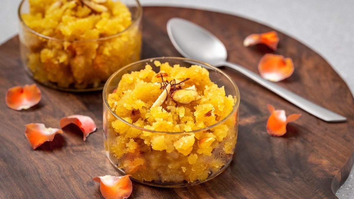 Moong Dal Halwa aus gelben Mungbohnen in Glasschalen auf Holzbrett. Daneben Blüten und Löffel. Draufsicht.