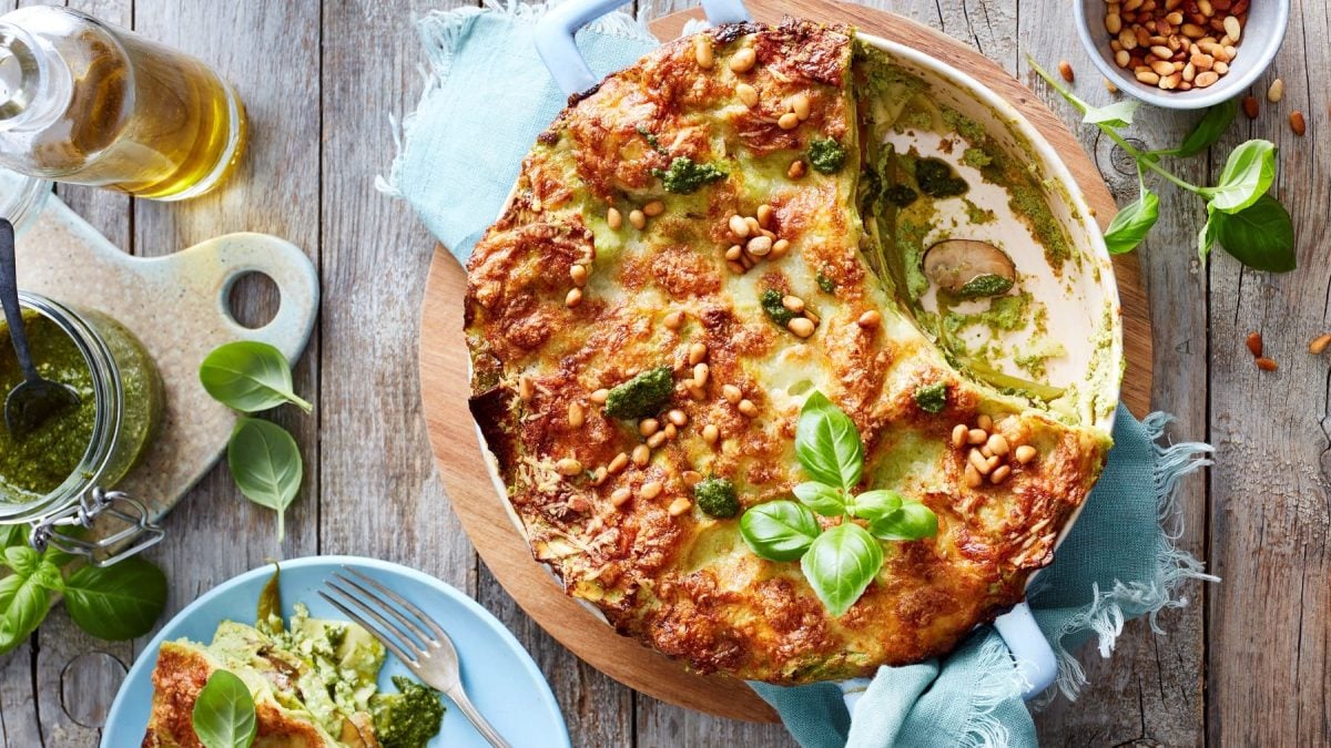 Runde Pesto-Lasagne in der Form neben verschiedenen Zutaten vor Holzhintergrund.