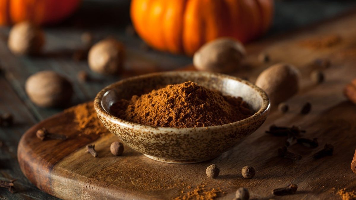 Selbst gemachtes Pumpkin Spice in Holzschale auf Holzbrett. Daneben Gewürze. Im Hintergrund Kürbisse. Frontalansicht.