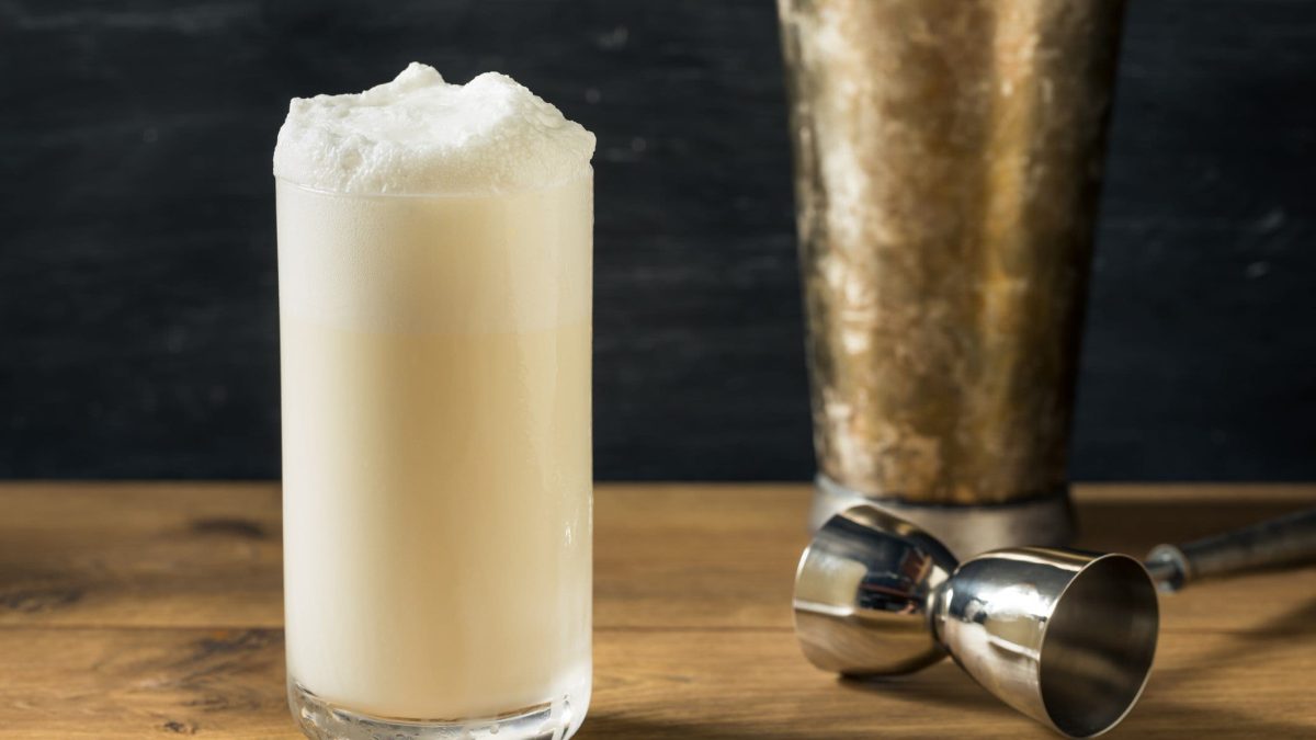 Ramos Gin Fizz mit Barmaß und Shaker auf Holz vor dunklem Hintergrund. Frontalansicht.