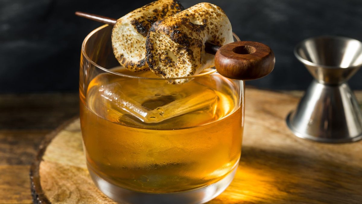 Rosemary Bourbon Candy Shock Cocktail mit Marshmallows auf Holzbrett auf Holztheke. Daneben Barmaß. Fronatlansicht.