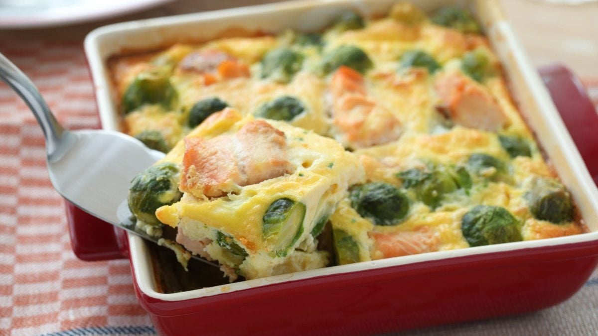 Rote Auflaufform mit Rosenkohl-Lachs-Gratin vor rot-weißem Hintergrund.