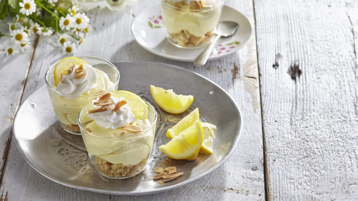 Schichtdessert mit Lemoncurd und Limoncello in Gläsern auf weißem Teller mit Zitronenspalten. Daneben Blumendekoration. Draufsicht.
