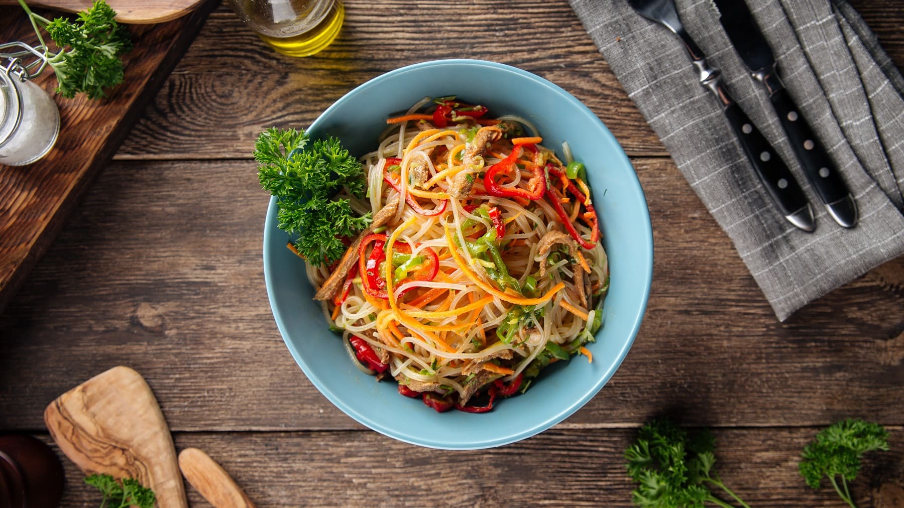 eine Schüssel schnelle asiatische Pasta mit Erdnusssauce und bunter Paprika sowie Petersilie in der Draufsicht, daneben Besteck.