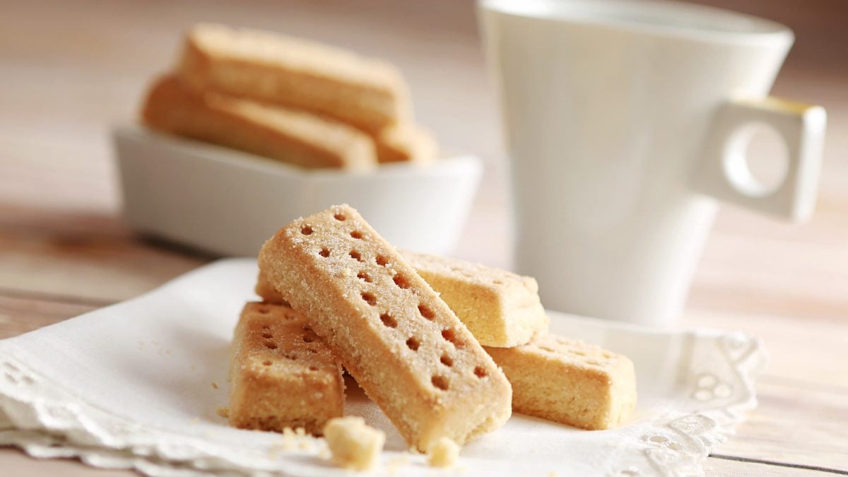 Schottisches Shortbread auf Stoffservierte und hellem Untergrund. Im Hintergrund eine weiße Tasse und eine Schale mit weiteren Shortbread. Frontalansicht.