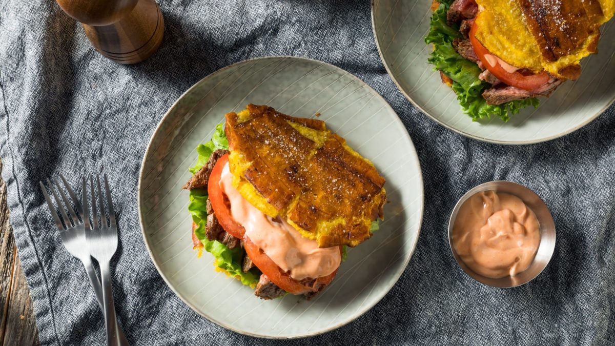 Zwei Tostones Burger auf zwei hellen Tellern neben einer Schale mit Sauce, einer Gabel und einer Gewürzmühle vor grauem Stoffhintergrund.
