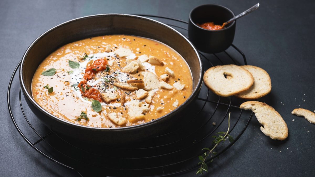 Vegane Paprika-Kartoffel-Suppe in einer schwarzen Schüssel neben Knoblauch-Brotchips und Ajvar vor schwarzem Hintergrund.