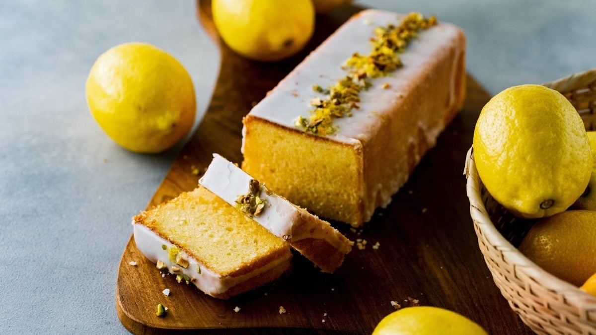 Ein angeschnittener veganer Kokoskuchen mit Guss und gehackten Pistazien dekoriert, drumherum liegen Zitronen.