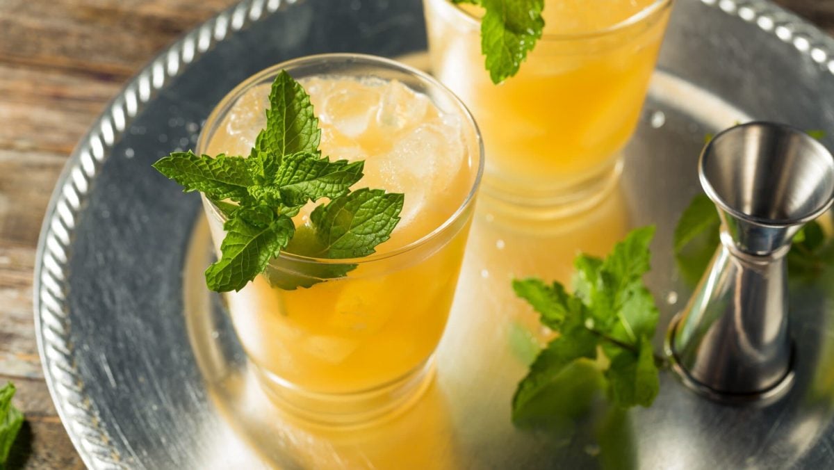 Whiskey Smash mit Minze im Glas auf Silbertablett. Daneben Minze und Barmaß. Draufsicht.
