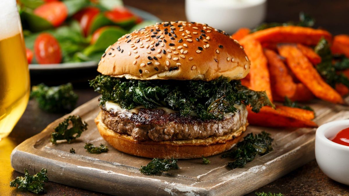 Wildschwein-Burger mit frittiertem Grünkohl auf Anrichtebrett. Im Hintergrund Süßkartoffelpommes, Dips, Salat und ein Glas Bier. Frontalansicht.