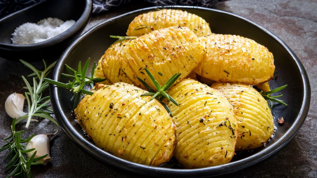Ein Teller zitronige Hasselback-Kartoffeln mit Rosmarin, daneben Rosmarinzweige und Knoblauchzehen.