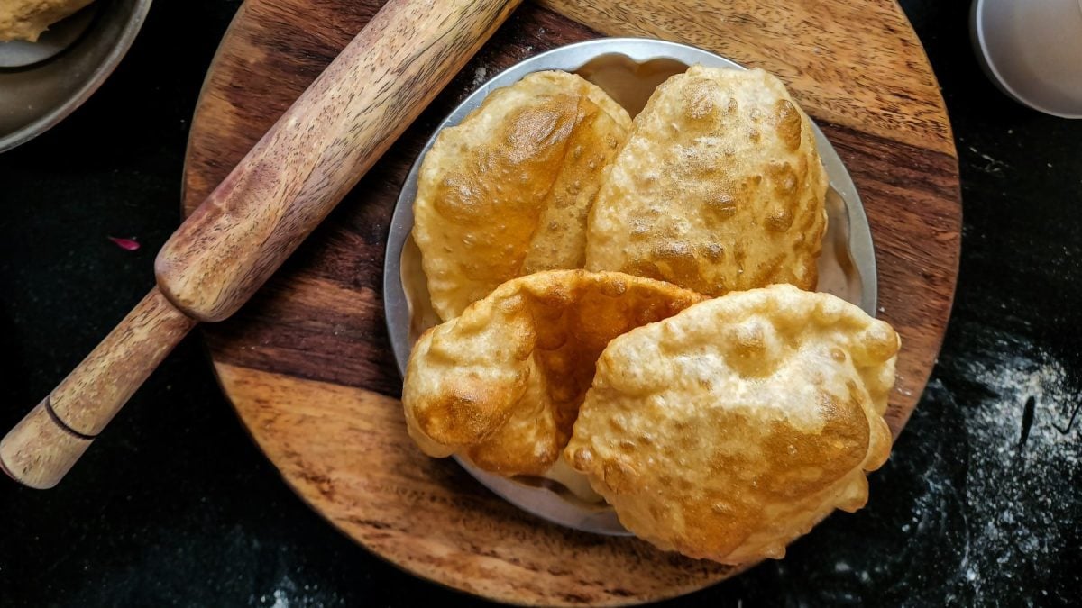 Bhatura selber machen: Mehrere Bhatura in einer Schüssel neben einem Nudelholz auf einem Holzbrett vor dunklem Hintergrund.