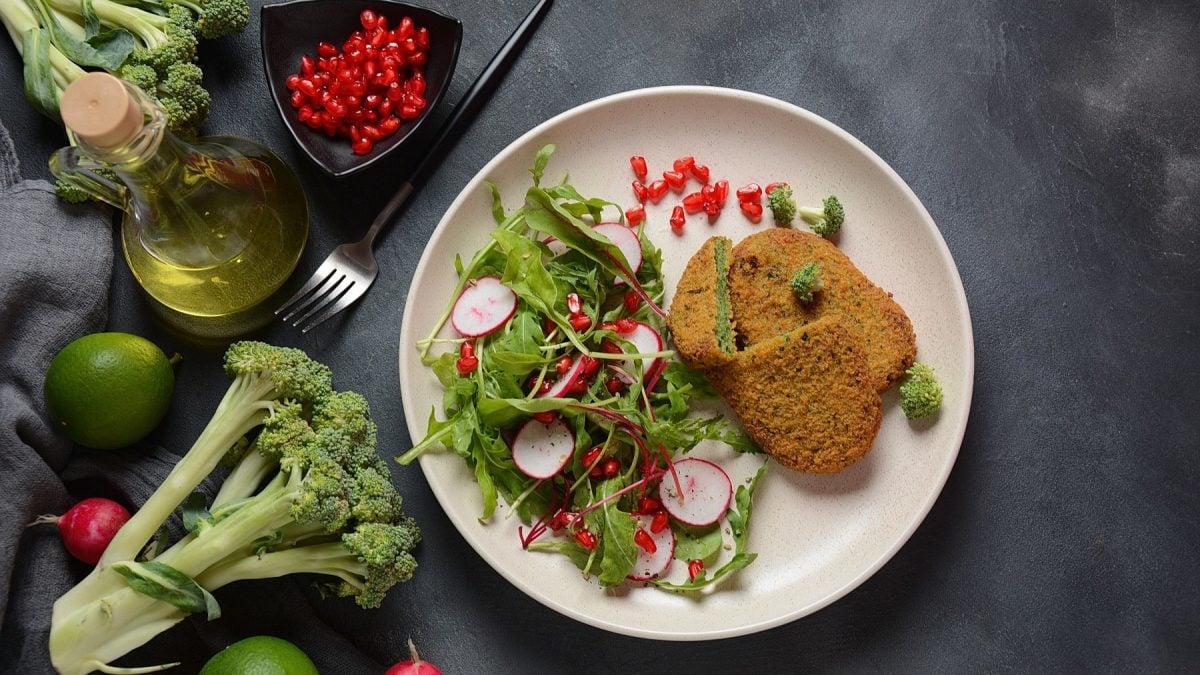 Brokkoli-Schnitzel mit Rucola-Radieschen-Salat, garniert mit frischer Vinaigrette und Granatapfelkernen, serviert auf einem weißen, flachen Teller auf einem steinernen Tisch.