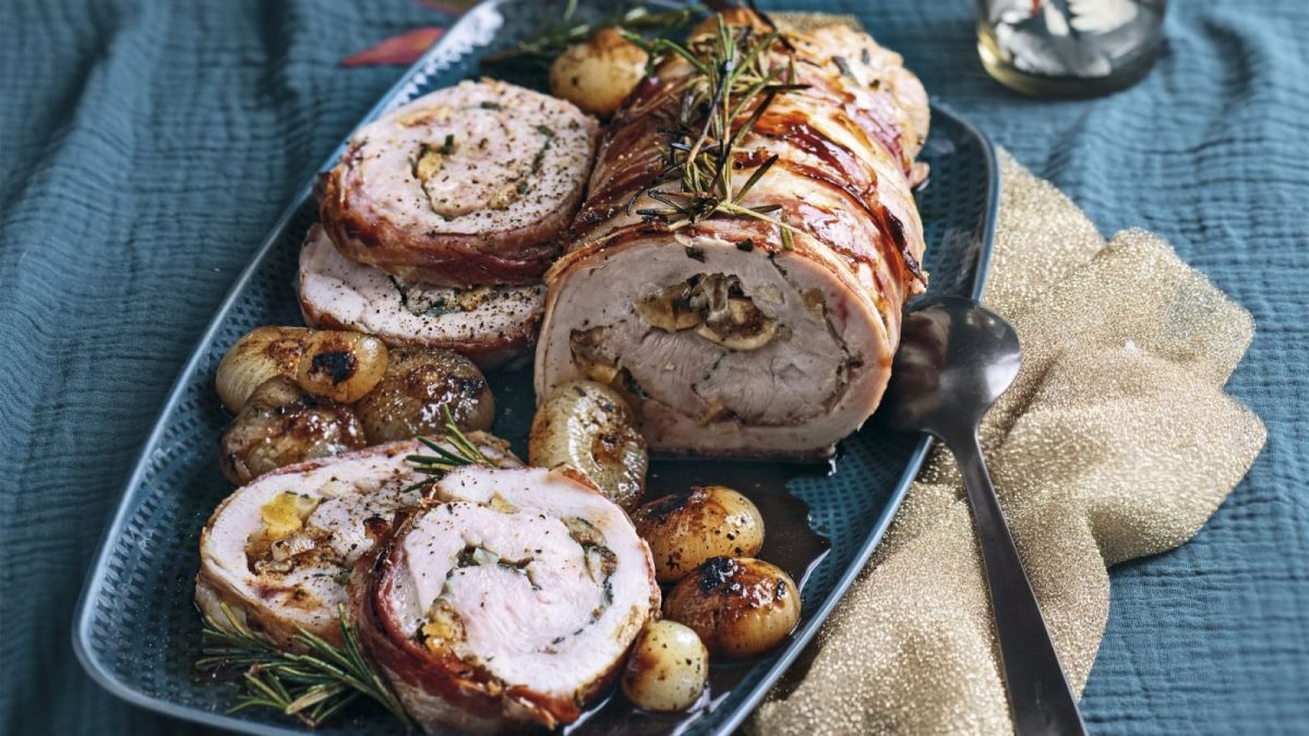 Gerollter Truthahnbraten gefüllt mit Feigen und Kräutern. Umwickelt mit Speck und garniert mit Rosmarin. Auf einem blauen Teller mit karamellisierten Zwiebeln serviert.