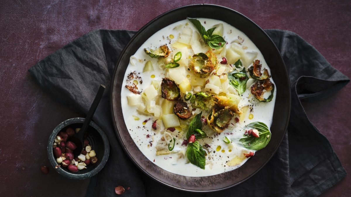 Kohlrabi-Kokos-Suppe mit paniertem Rosenkohl, Erdnüssen, Chiliflocken und frischem Thaibasilikum, serviert in einer dunklen Keramikschale auf dunklem Untergrund.