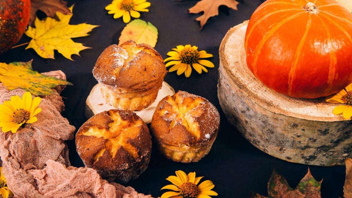 Drei herzhafte Kürbismuffins vor dunklem Hintergrund mit Herbstblättern, einem Kürbis und gelben Blüten als Deko.